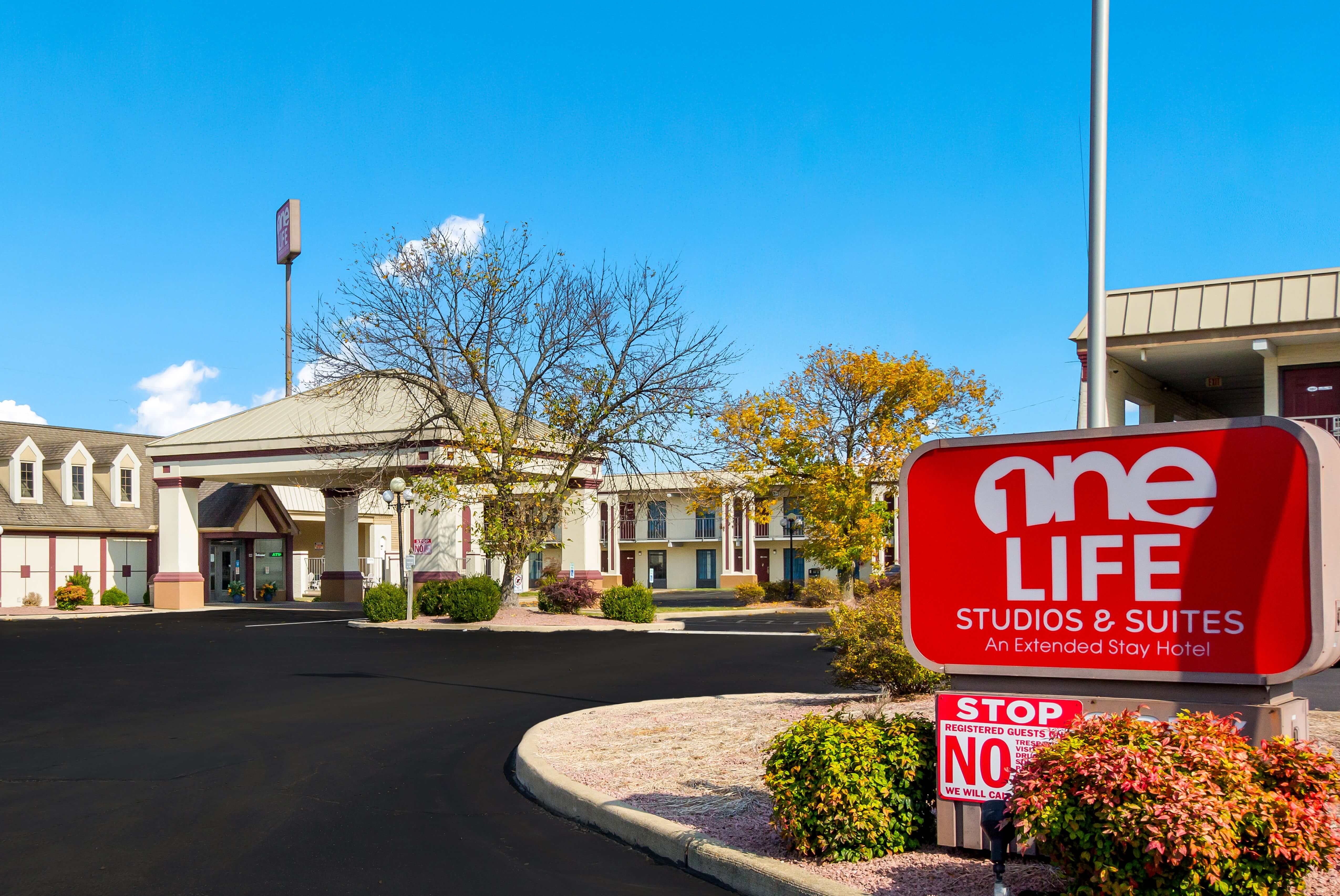 A red sign with white text in front of a white building with a parking lot and trees.