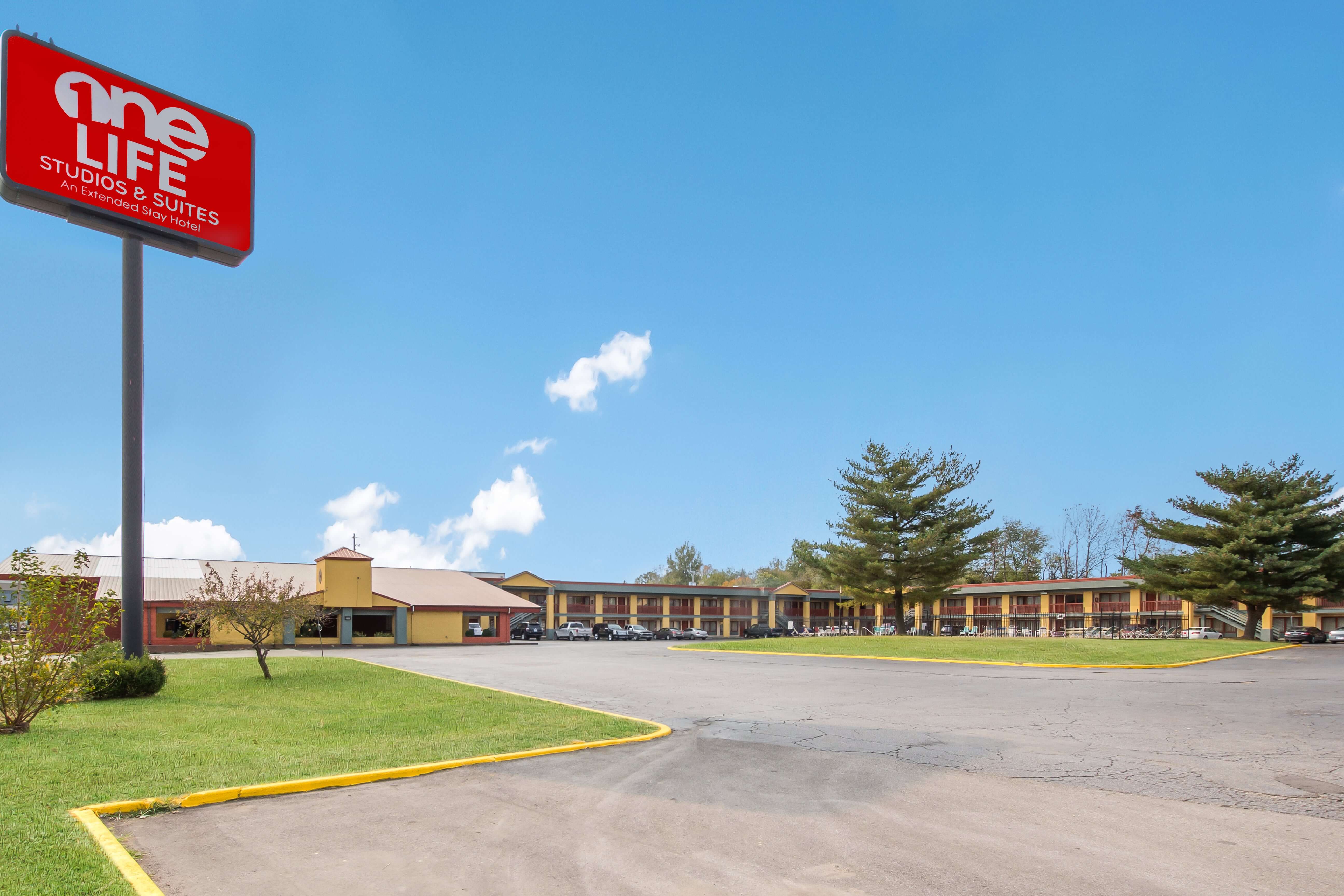 A red sign with white text stands in front of a yellow hotel building with a parking lot.