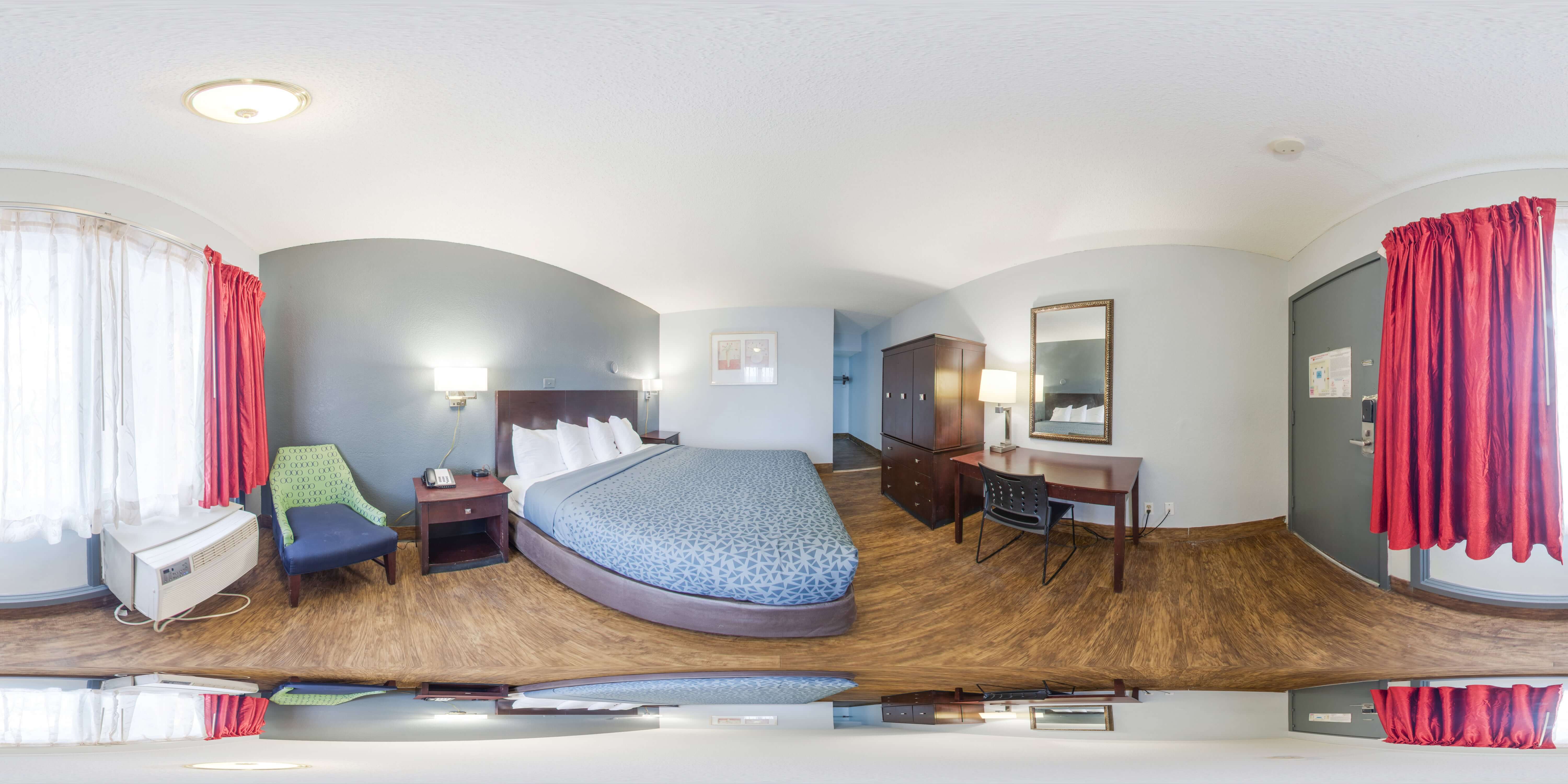 A wide-angle view of a hotel room with a bed, desk, chair, and window.