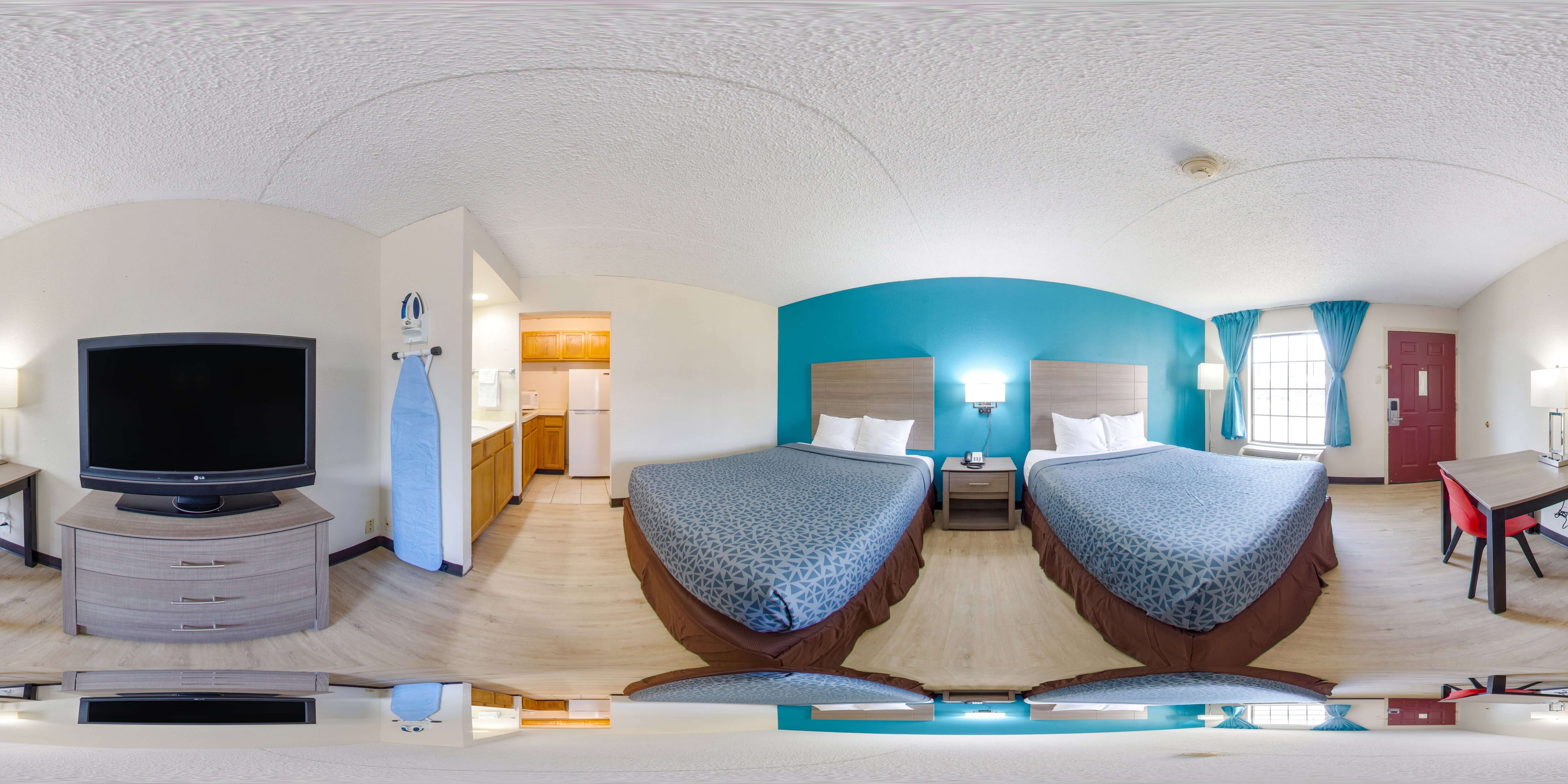 A wide-angle view of a hotel room with two beds, a desk, a television, and a kitchenette.