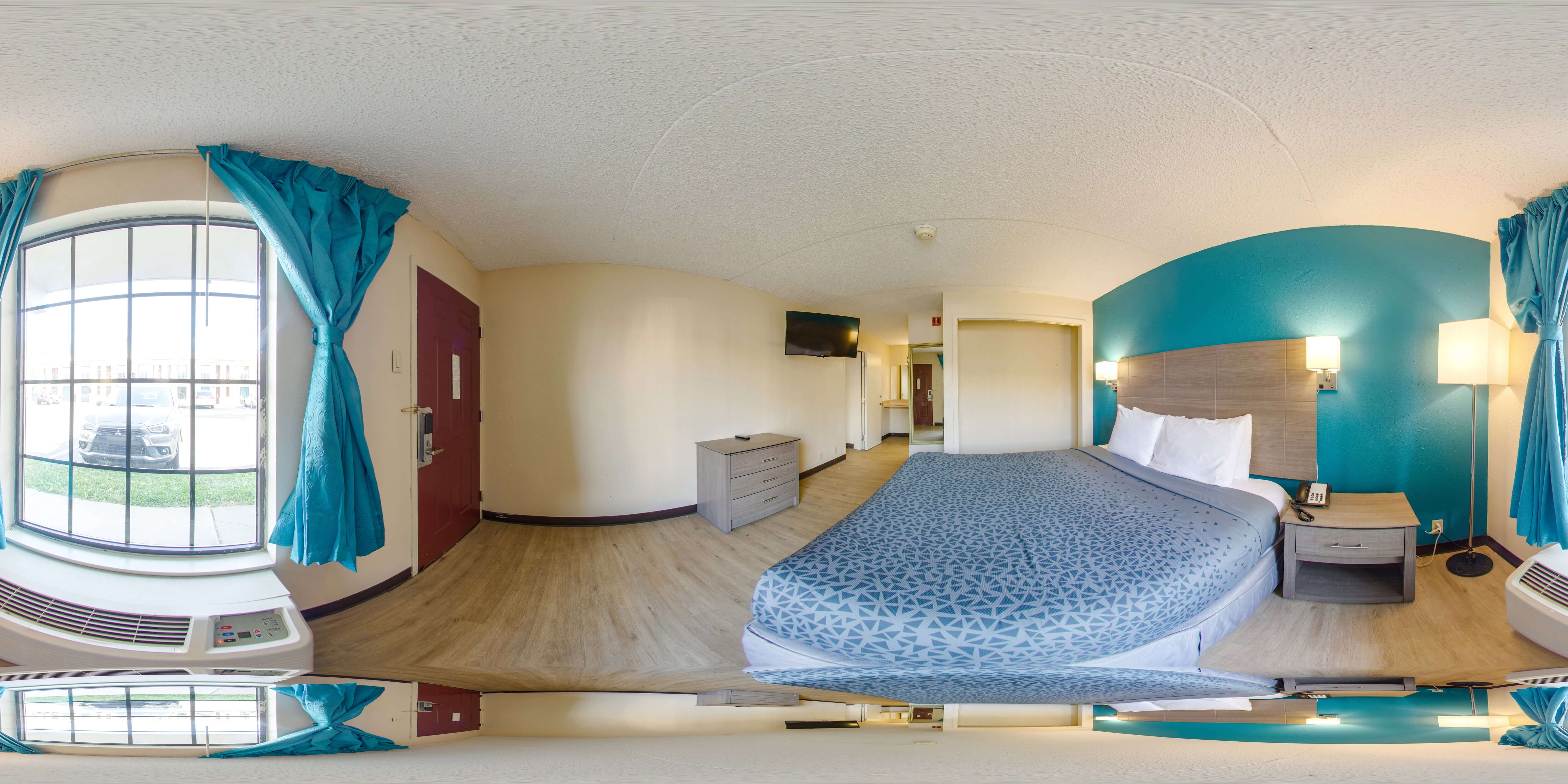 A wide-angle view of a hotel room with a king-sized bed, large window, and flat-screen TV.