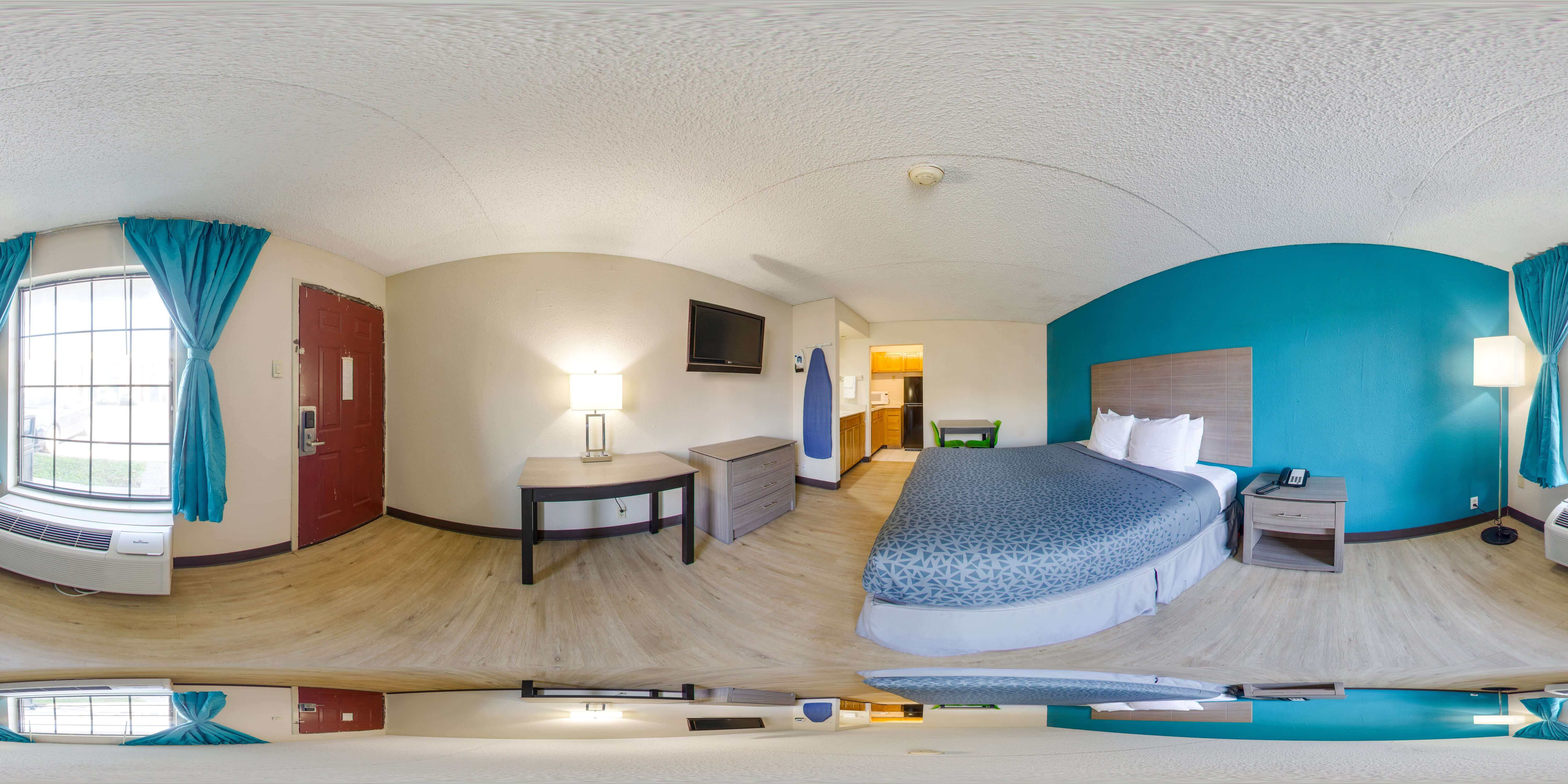 A wide-angle view of a hotel room with a bed, desk, kitchenette, and TV